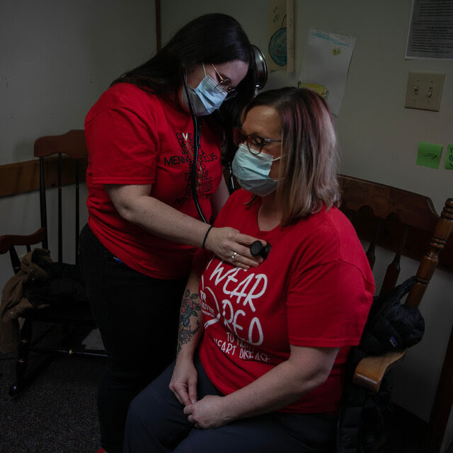 Dr. Joanna Bailey wears a red T-shirt and listens to the heart of a patient, who is also wearing red, with a stethoscope in an exam room of her clinic. Both of them wear surgical masks.