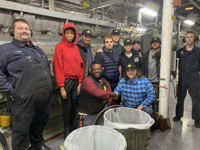 Group of people in a ferry engine room, wearing ear protection and posing for the photo