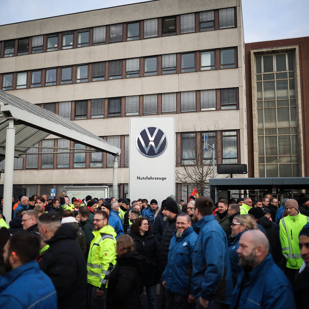 Employees wearing winter jackets stand outside of a large business office with a Volkswagen sign.