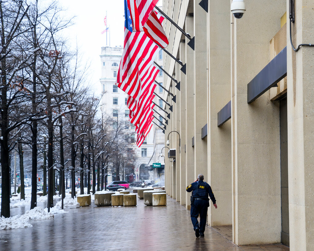 A man wearing a vest with “FBI” on it outside a large building with American flags overhead.