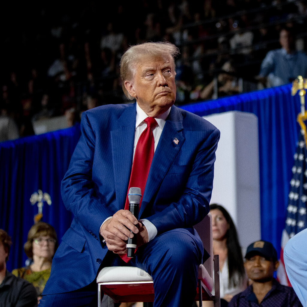 A man wearing a blue suit and red tie sits on a chair holding a microphone. 