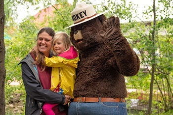 A smiling woman holds a little girl in her arms, standing next to someone in a Smokey Bear costume