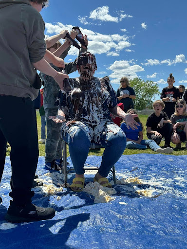 A person on a chair outdoors, covered in whipped cream and chocolate syrup, with people pouring more on them. Set on a blue tarp with onlookers, under a clear sky with scattered clouds.