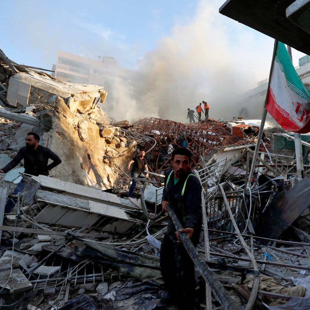 A man stands in front of a destroyed building with the Iranian flag in the background.