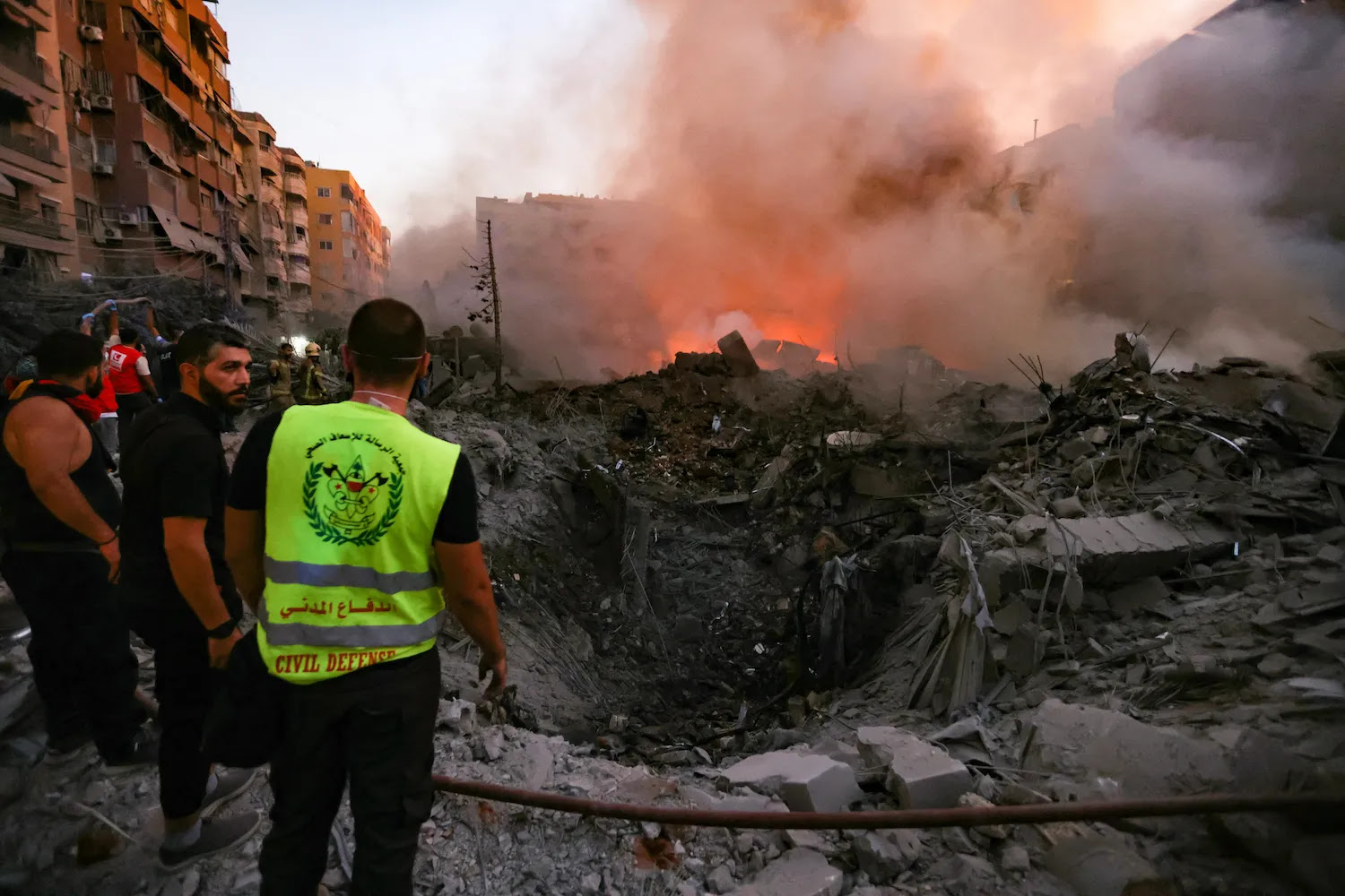 People gather on a street in Beirut around the rubble of a hole created by an Israeli airstrike. Smoke billows up from the impact site, and some orange flames are visible in it. Battered midrise buildings loom on either side.