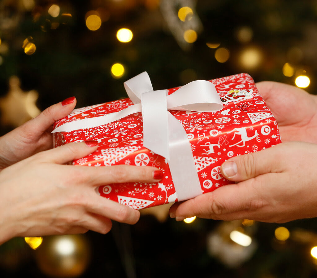 Two sets of hands hold a gift, which is wrapped in red patterned paper and a white bow.