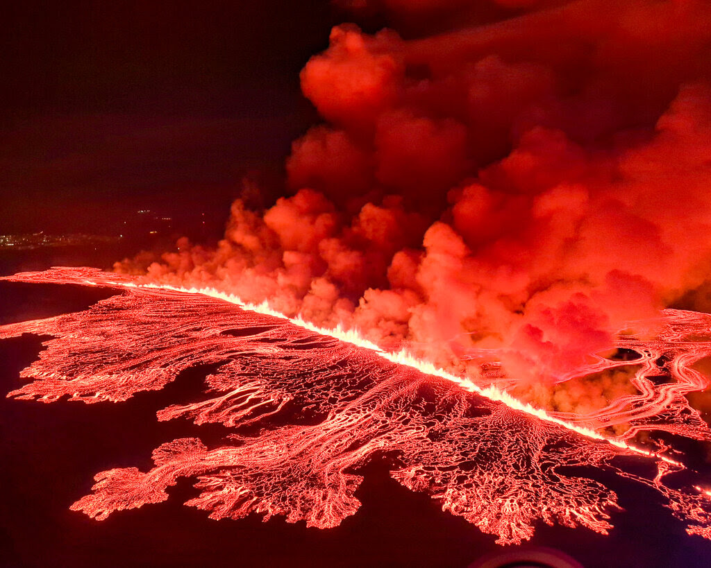 Reddish-orange lava flows and smoke billows during a volcanic eruption.