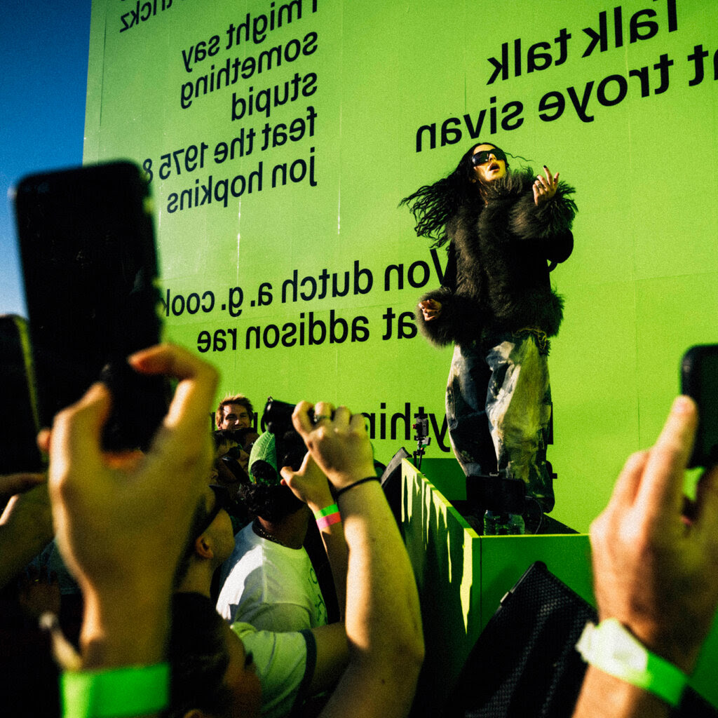 A woman in a dark fur coat and black sunglasses stands on a bright green D.J. booth. A young crowd watches with their phones out.