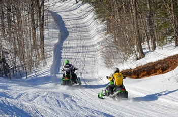 Two people in winter gear ride green and black snowmobiles down a wide, gently sloping, well-groomed trail surrounded by trees