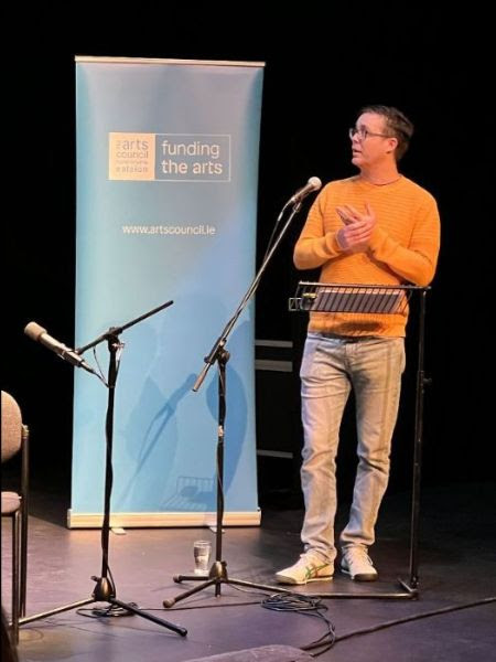 Dr Philip Finn stands at a podium, speaking in front of a blue Arts Council pop-up with the Arts Council logo. He wears a mustard jumper, grey jeans, and glasses.
