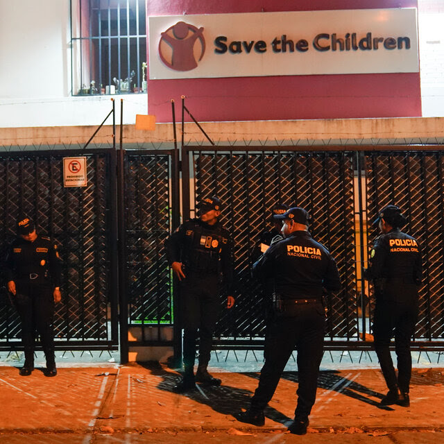 Police officers in uniform stand outside a building that has a fence in front of it with a sign that reads Save the Children.