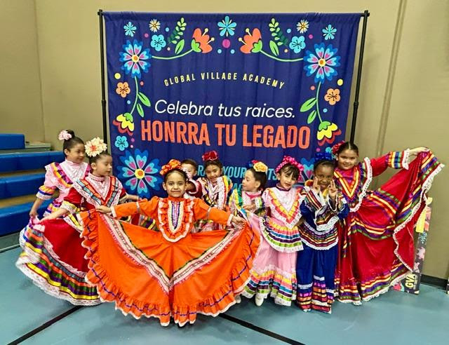 A group of young girls dressed in vibrant traditional dresses posing in front of a Global Village Academy banner that reads Celebra tus raices Honra tu legado smiles and colorful floral decorations behind them