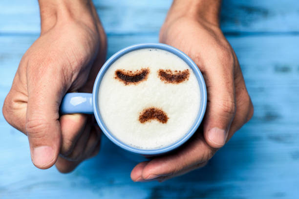 man with a cup of cappuccino with a sad face high-angle shot of a young caucasian man with a blue cup of cappuccino with a sad face drawn with cocoa powder on the milk foam, on a blue rustic table blue monday stock pictures, royalty-free photos & images