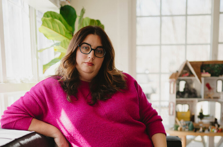 Virginia Sole-Smith, wearing a pink sweater and glasses, posing by a window with sunshine coming through.