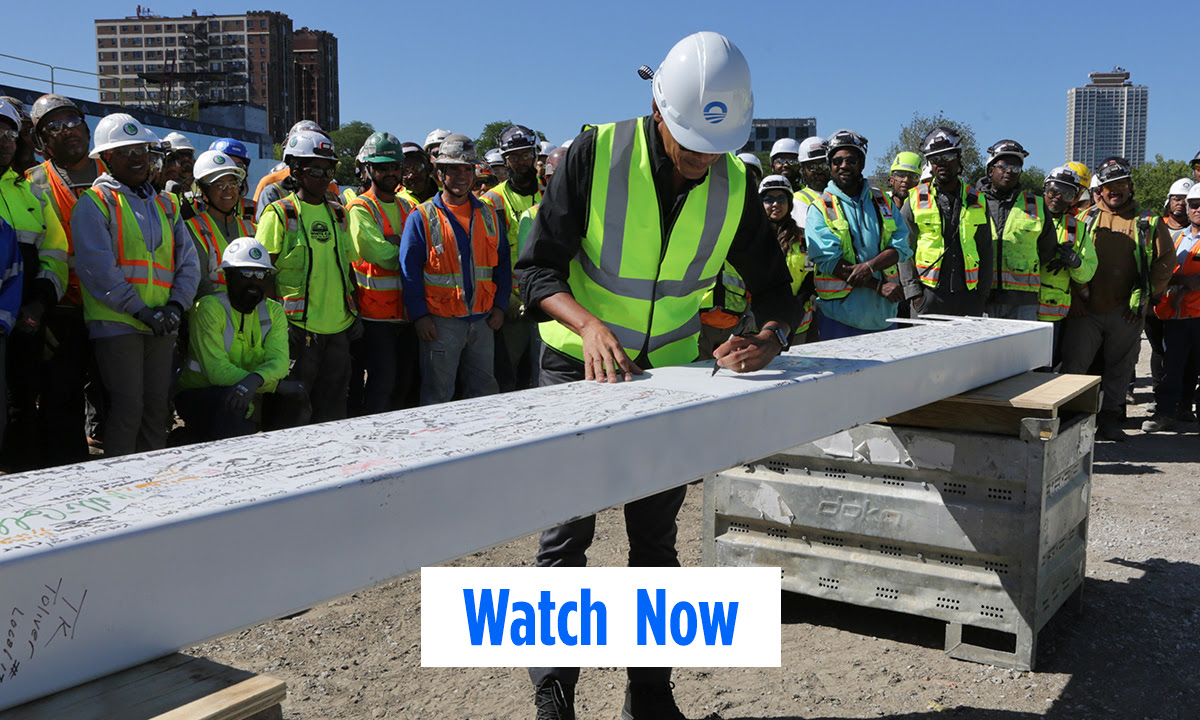 President Obama is wearing construction gear and signing a beam with a sharpie while construction workers look on