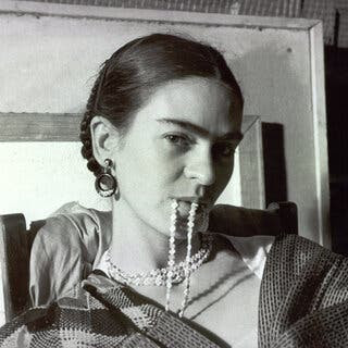 In a black-and-white image, Frida Kahlo looks temptingly at the camera. In her mouth is part of a string of beads that’s wrapped three times around her neck.