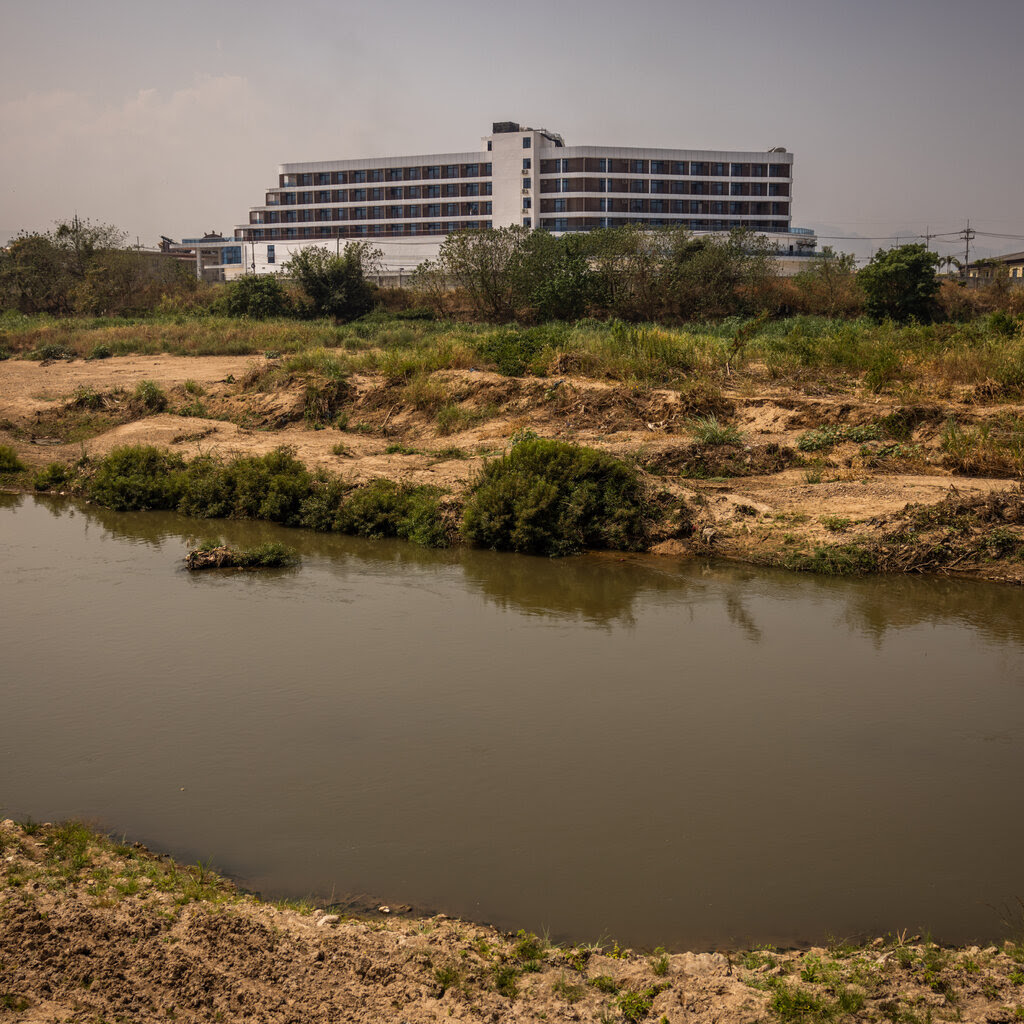 A new mid-rise building stands out amid a mostly barren landscape as seen from across a narrow river.