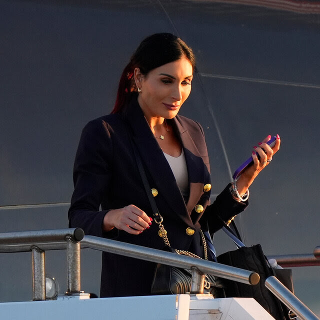 Laura Loomer, carrying luggage and holding a smartphone in one hand, exits an airplane onto a set of stairs.