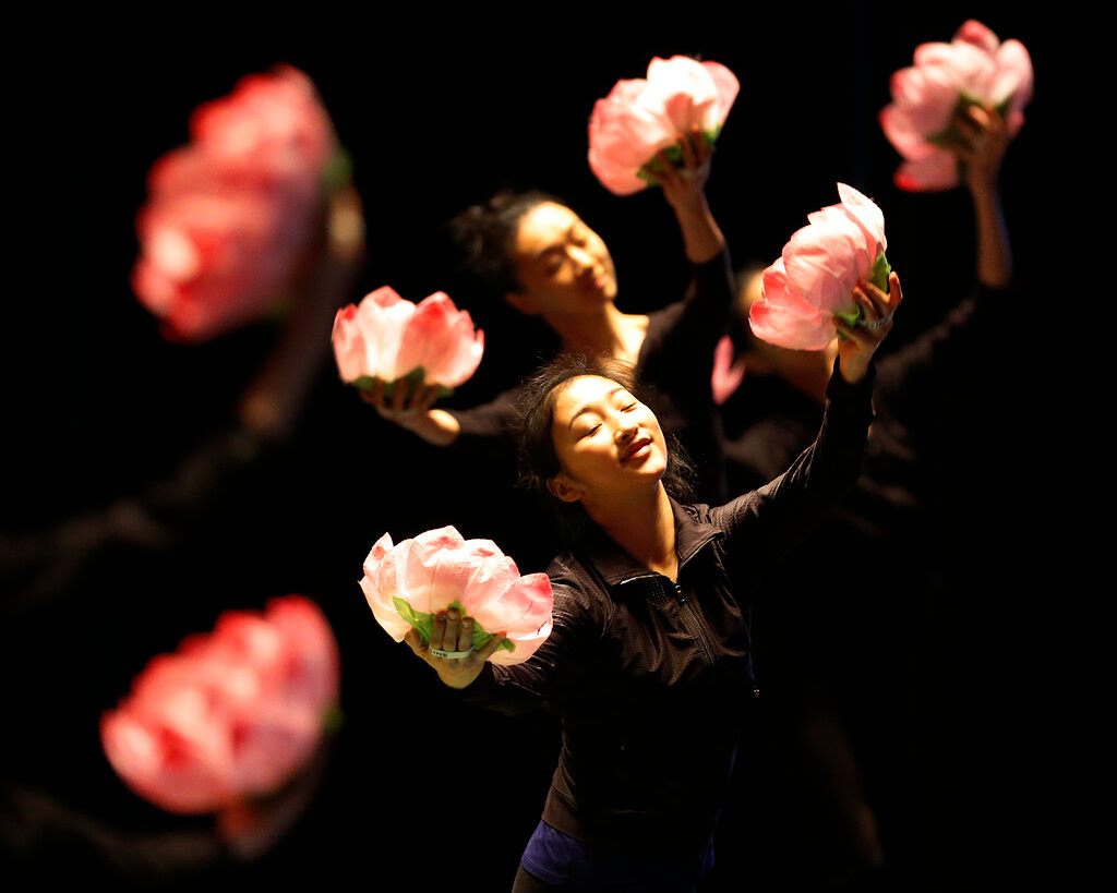 Dancers wearing black hold up pink paper flowers against a black background. 