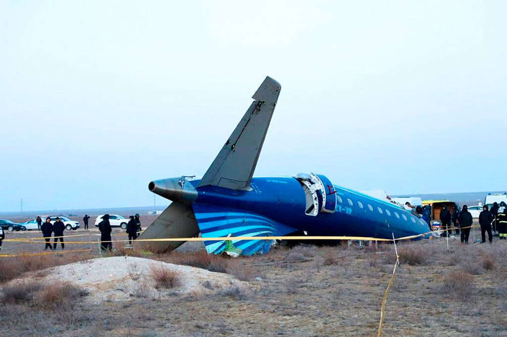 The rear half of an airplane lying overturned on scrubland, with people and vehicles nearby.