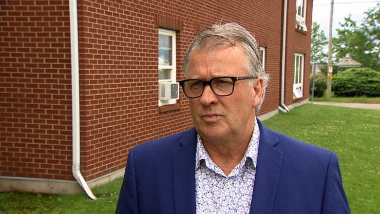 A man with a flowered button-up shift and a blue blazer stands outside a brick building.