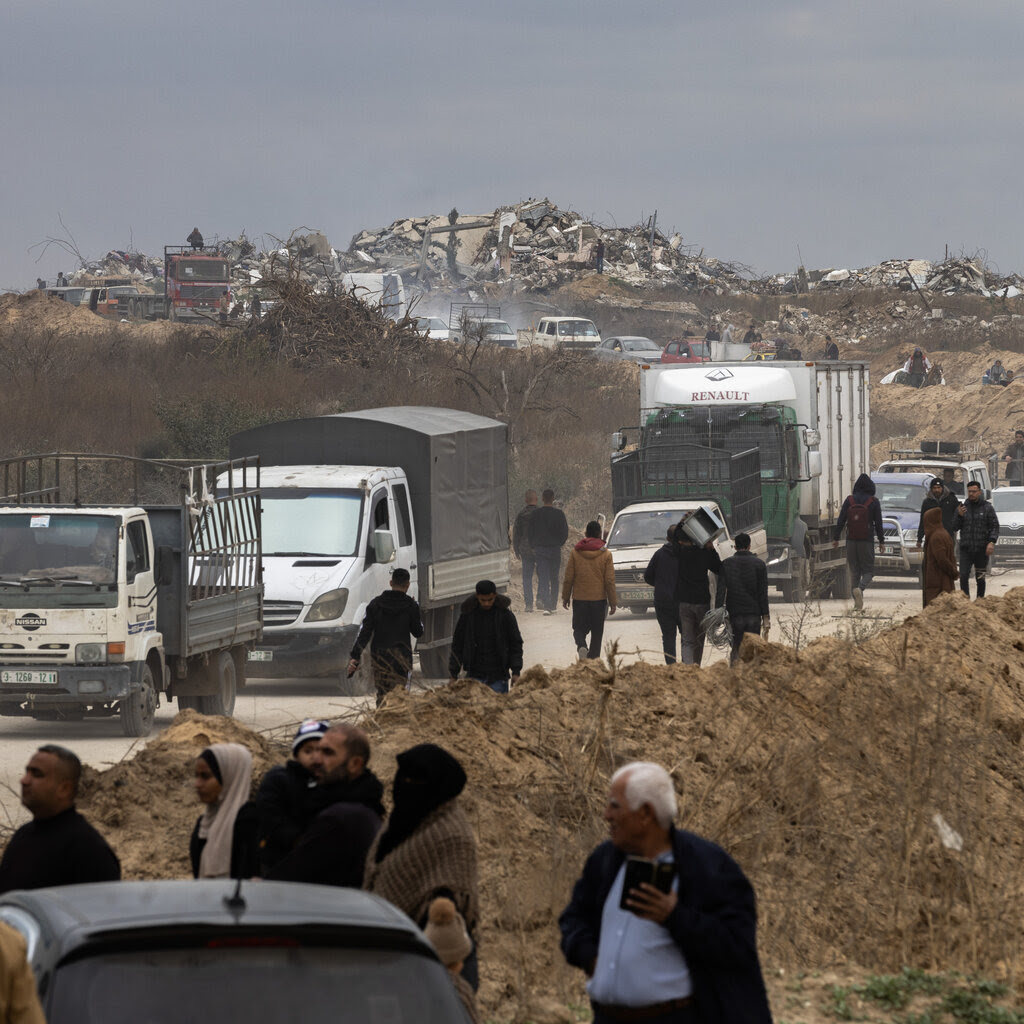 A line of cars and trucks is seen on a road. Rubble is in the distance.