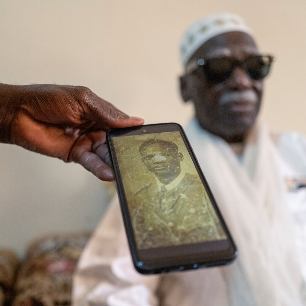 An old portrait of a man displayed on a phone, with a man in white sitting in the background.