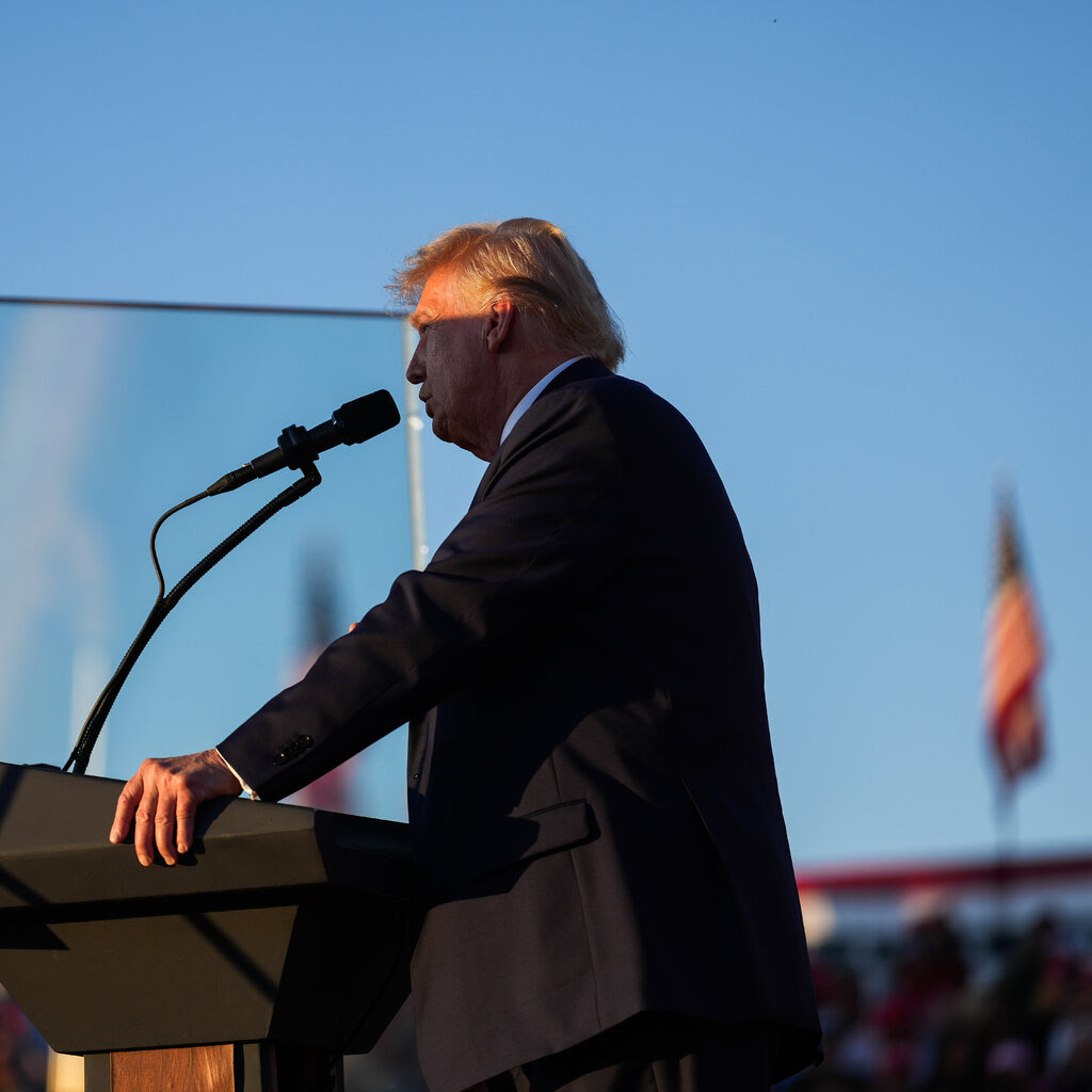 Donald Trump stands at a podium. A shadow is cast over part of his face.