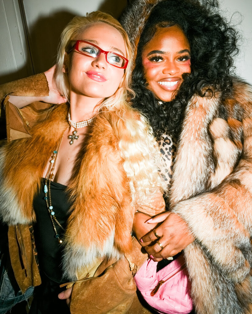 Two young women pose arm-in-arm, wearing vintage-look fur jackets.