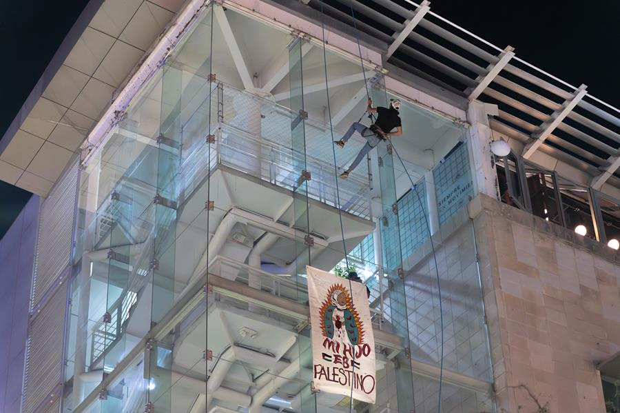 A banner of the Virgin of Guadalupe with the words "My hijo es Palestino" (English translation: My son is Palestinian). The banner is hanging over the glass on the side of a building and there is a person rappelling down the side of the building.