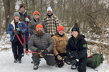 small group of men and women in winter gear and flannel, some holding trimming tools, on a snow-covered road in forest