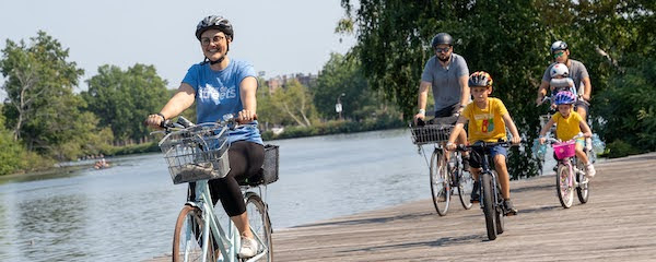 LivableStreets Executive Director Stacy
Thompson on the family ride at tour de streets 2023. Riding her light
blue bike with a basket in front, Stacy is wearing a light blue Tour
de Streets t shirt and a grey helmet. Behind her are a few adult and
child bike riders; next to the boardwalk where they are biking is the
Charles River