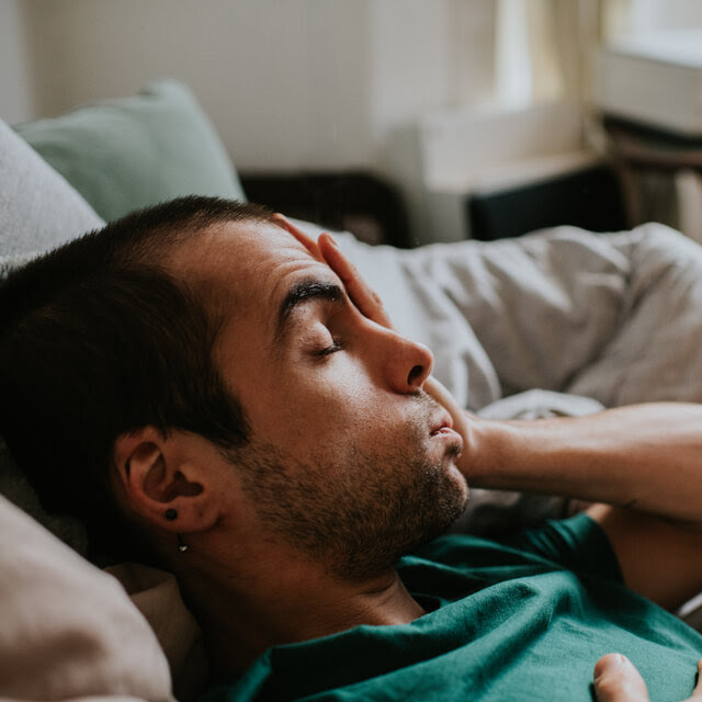 A man lies back on a couch among pillows. He rubs his eyes and face.