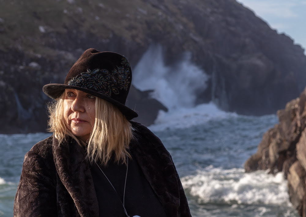 Poet Dairena Ní Chinnéide, in dark attire, looks to her right, while in the background choppy seas crash against rocks.