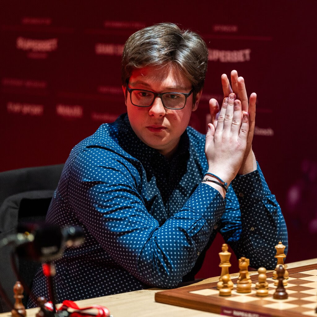 A man in glasses and a blue shirt sits in front of a chess board.