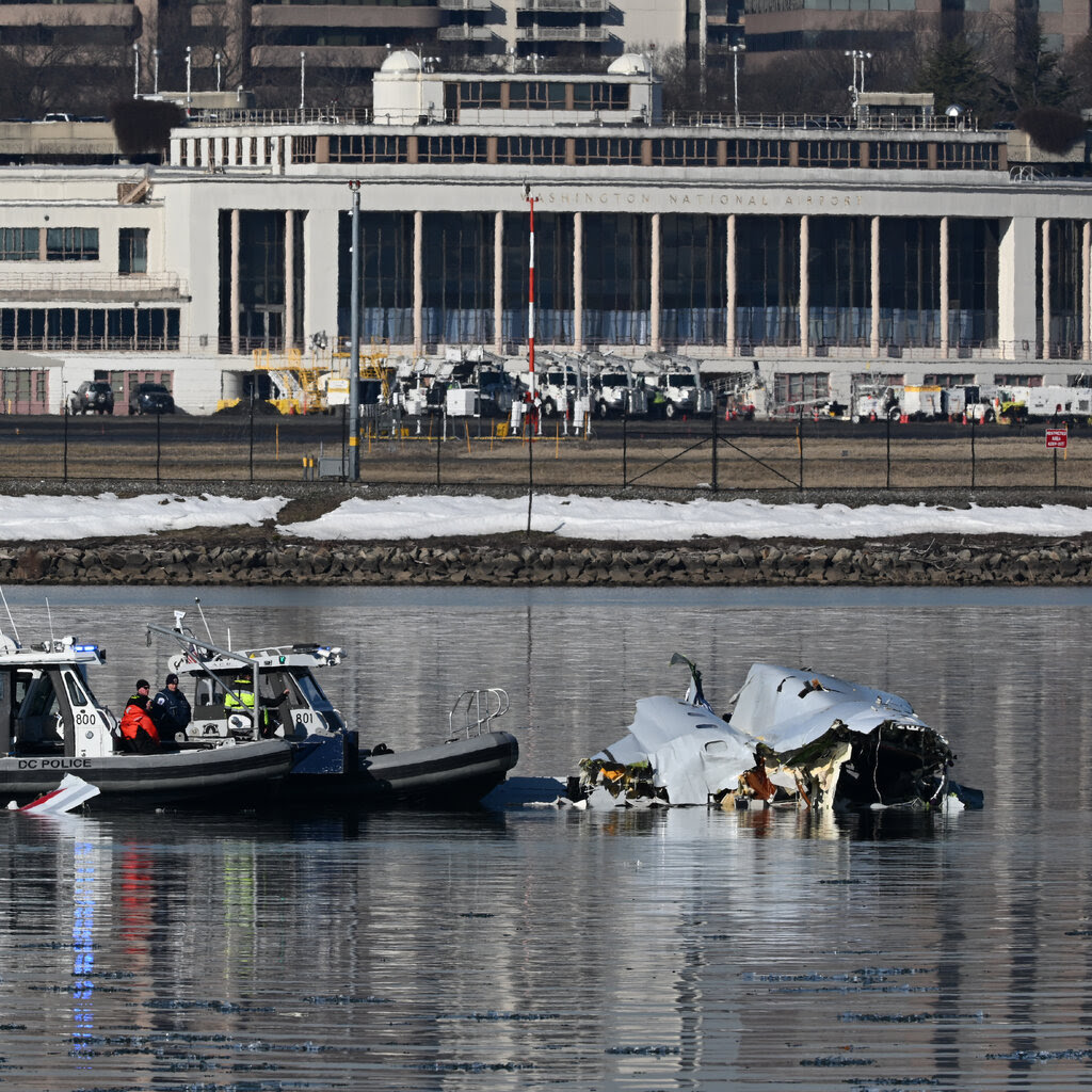 Plane wreckage floats in a river. Two police boats sit next to the wreckage. In the background are multiple buildings.