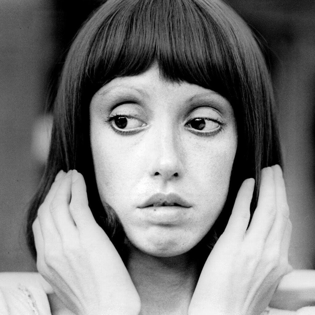 Shelley Duvall in a black-and-white portrait from the 1970s, holding her hands up to the sides of her head.