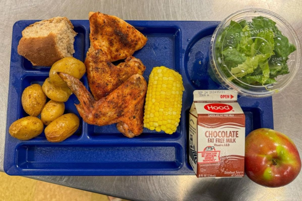 Lunch tray with small potatoes, a roll, chicken, half an ear of corn, chocolate milk, salad, and an apple