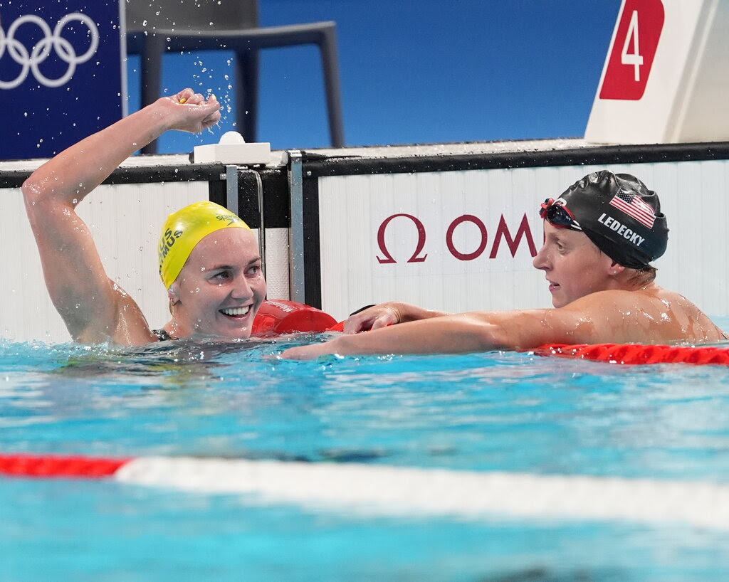 Ariarne Titmus raising her arm and smiling in the pool lane next to Katie Ledecky.