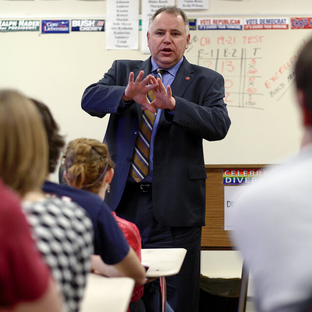 Tim Walz, wearing a suit and tie, gestures with his hands as speaks to a class of students.