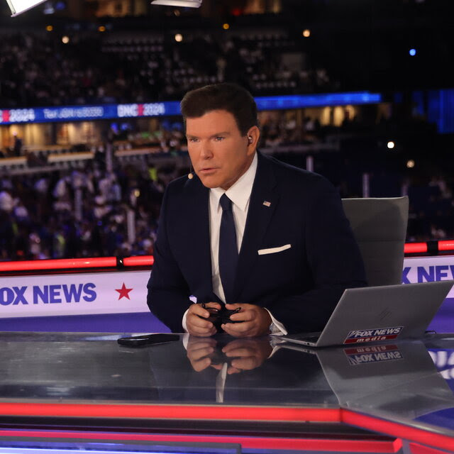 Bret Baier sentado detrás de un escritorio de Fox News habilitado dentro de un estadio.