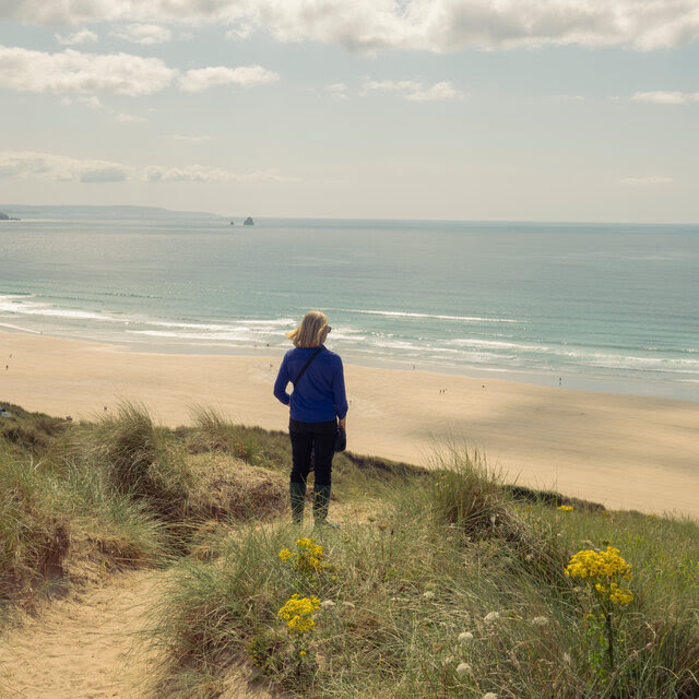 A woman turning her back to the camera on the dunes of a beach. The water is visible in the background and the dunes are grassy. There are flowers. 