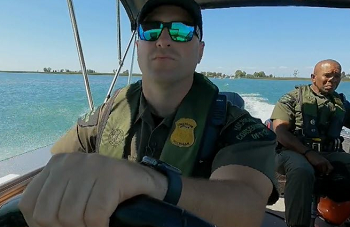 a man in sunglasses and conservation officer uniform drives a boat on open blue water, with another man in uniform sitting behind him