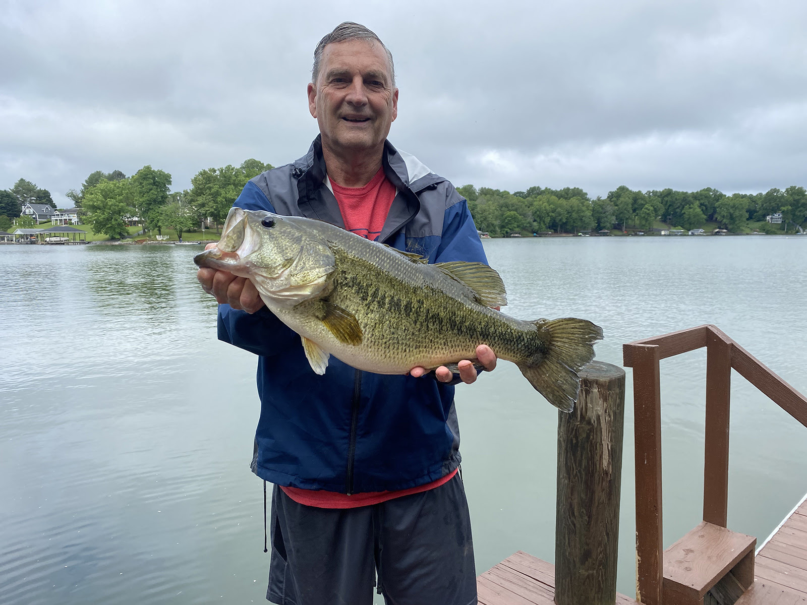 A photo of a man standing on a dock in front of a lake holding a very large largemouth bass.