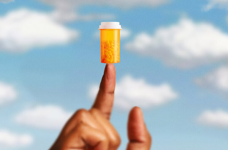 A photo illustration of a hand and index finger pointed up. The background is a blue sky with white clouds. On top of the index finger is an orange prescription bottle with pills in it. 