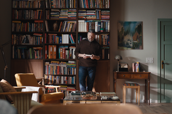 Man in brown jumper stands in front of a very full bookcase 