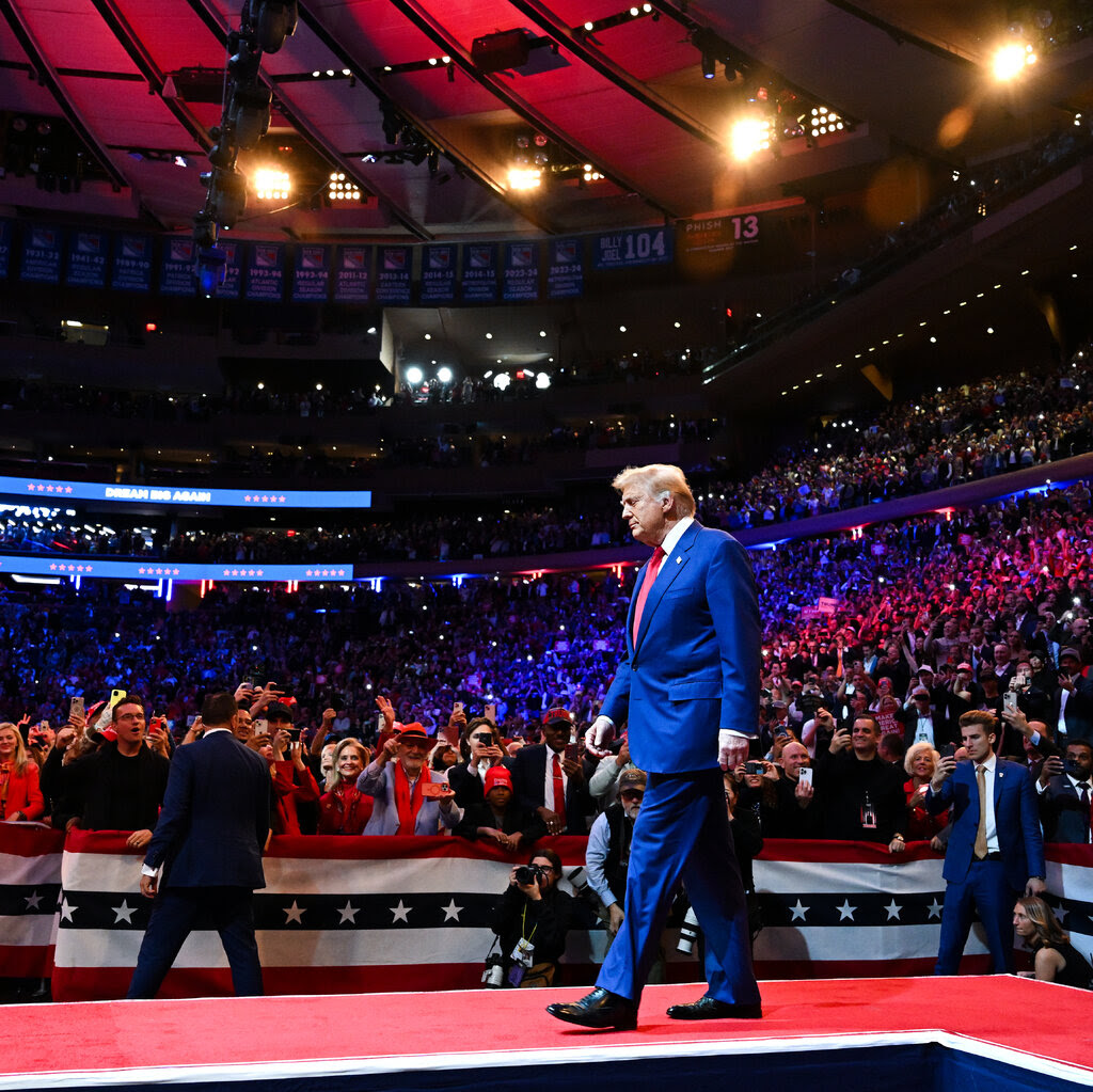 Donald Trump walks across a stage before a crowd at a giant arena. 