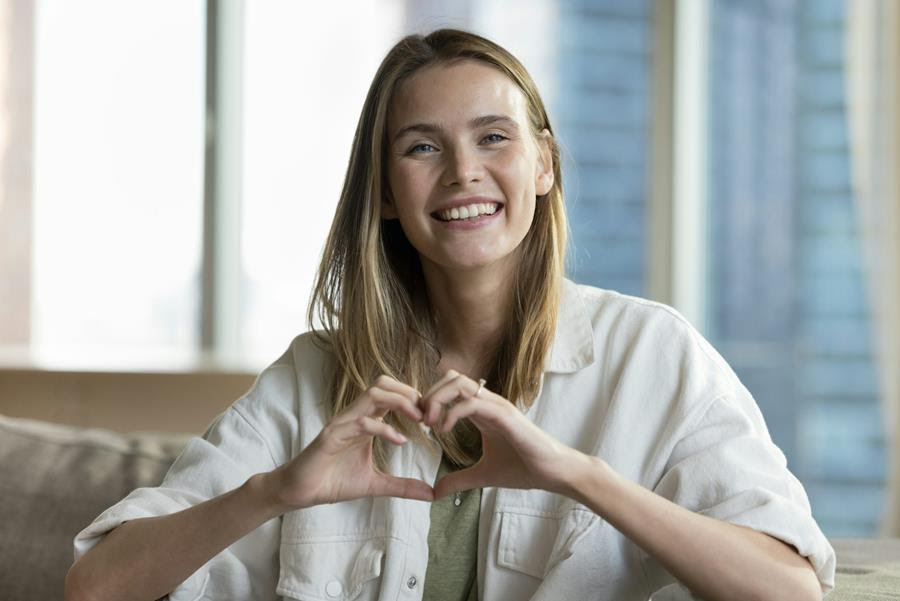 A smiling young woman looks at the camera, forming a heart shape with her hands.