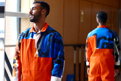 A photograph of a model wearing a jumpsuit with fabric designed by Zephir Liddell. The model is standing their back to a mirror in a dance studio, making the front and back of the jumpsuit visible. The jumpsuit is bright orange with a printed yoke and sleeves. The print is blue with abstract black lines. 