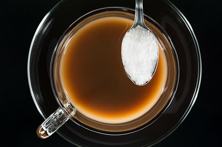 Artificial non-sugar sweeteners shown on a spoon hovering over a glass cup and saucer of tea